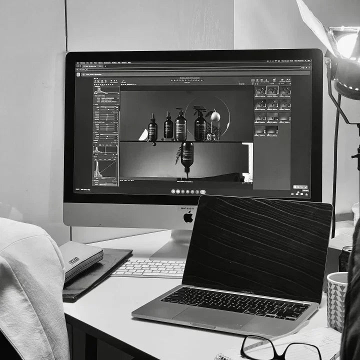 Black and white photo of a desktop with iMac displaying product photo editing, MacBook, and studio light.