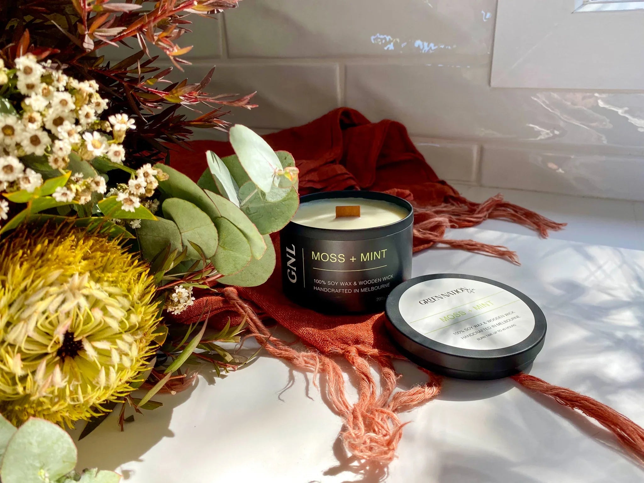 Moss + Mint soy candle in black tin with wooden wick beside floral bouquet on white counter