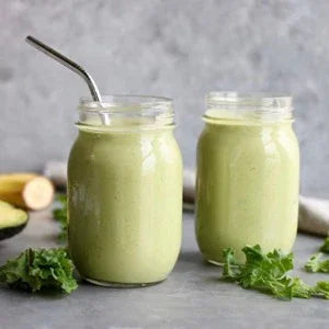 Two green smoothies in mason jars with a metal straw, kale and banana visible on a gray surface