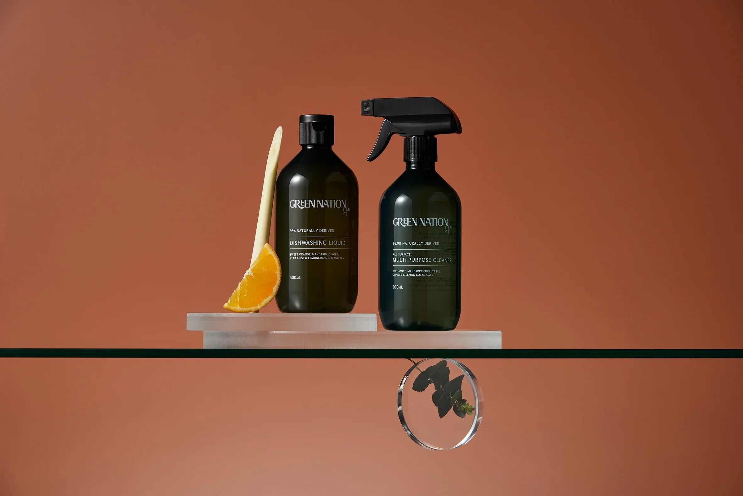 Green Nation dishwashing liquid and multi-purpose cleaner bottles with orange slice and lemongrass on glass shelf, brown background
