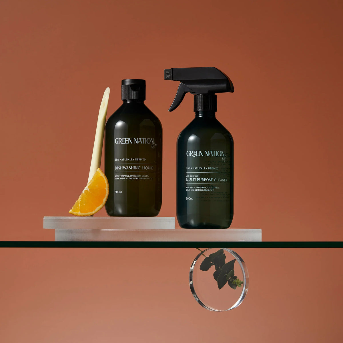 Green Nation dishwashing liquid and multi purpose cleaner bottles with orange slice and lemongrass on glass shelf against brown background